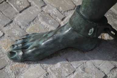 Bronzeskulptur eines Fußes mit einer Kette am Knöchel, auf Steinen.