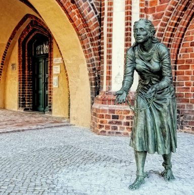 Bronzeskulptur einer Frau in Ketten gefesselt vor einem historischen Backsteingebäude.